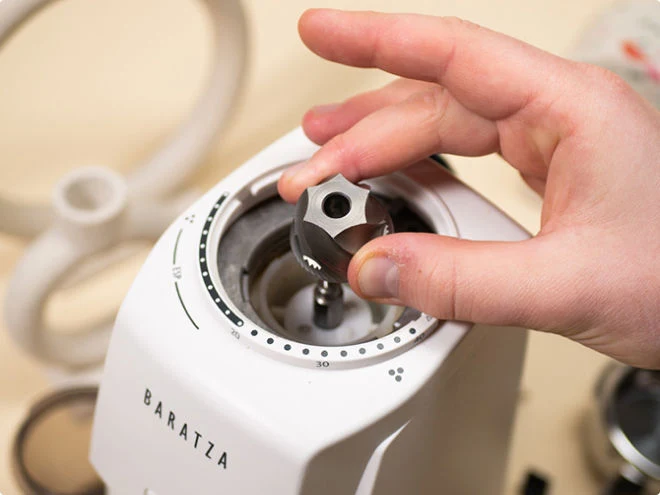 A hand lifts the central cone burr out of a white Baratza grinder